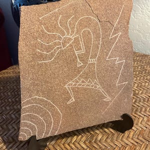 May include: A rectangular, reddish-brown stone with a white etched Kokopelli figure, lightning bolt, and sun design. The stone is displayed on a dark brown stand, showcasing a Southwestern theme. The artwork is a unique decorative piece.
