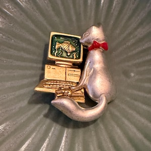 May include: A silver-toned cat brooch with a red bow tie, positioned next to a gold-toned computer. The computer screen displays a green background with a gold fish graphic. The cat is depicted with one paw resting on the computer keyboard.