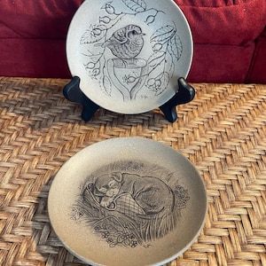 May include: Two small ceramic plates with a brown glaze. The top plate features a black and white illustration of a bird perched on a branch with berries. The bottom plate features a black and white illustration of a sleeping puppy.