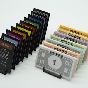 Könnte beinhalten: Ein Satz Monopoly-Spielkarten und Geld in verschiedenen Farben, darunter Schwarz, Blau, Grün, Gelb, Orange und Lila. Die Karten sind in einer Reihe angeordnet, und das Geld ist in einem Stapel gestapelt. Das Geld ist in Stückelungen von 1 €, 5 €, 10 €, 20 €, 50 € und 100 €.