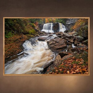 May include: A scenic photograph of a waterfall cascading over rocks in a forest setting. The water is flowing over the rocks and creating a white, foamy effect. The trees are mostly green with some yellow and orange leaves. The scene is bathed in a soft, natural light.