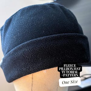 May include: A dark navy blue fleece pillbox hat. The hat is displayed on a mannequin head. The image includes a white and black graphic with the text "FLEECE PILLBOX HAT TUTORIAL + PATTERN" and "One Size".
