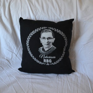 May include: Black throw pillow with a white image of Ruth Bader Ginsburg, a Supreme Court Justice, with the text "Notorious RBG" in a white wreath.
