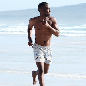 May include: A man runs on a beach wearing white swimming trunks with a blue and white pattern. The swimming trunks have the word "travel" printed on them.