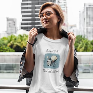 Puede incluir: Camiseta blanca con un gráfico de una cámara vintage y un globo aerostático, con el texto "Marked Safe From Spy Balloon". La camiseta la lleva una persona con gafas y una chaqueta vaquera.