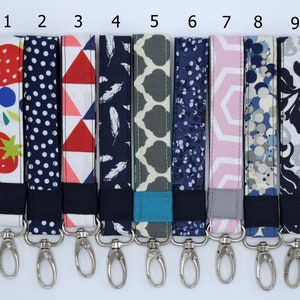 May include: Nine colorful fabric keychains with silver swivel clips. The keychains feature various patterns, including strawberries, polka dots, geometric shapes, feathers, and floral designs.