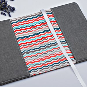 May include: A gray fabric book cover with a white ribbon bookmark. The inside of the cover is lined with a white fabric with a colorful wavy pattern in red, blue, turquoise, and gray.