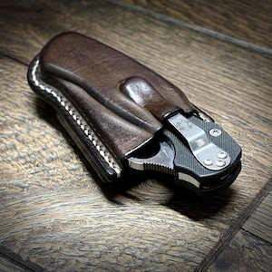 May include: A dark brown leather sheath containing a black folding pocket knife. The sheath features visible white stitching along the edge. The knife handle is black with a silver metal pocket clip attached by three small screws.