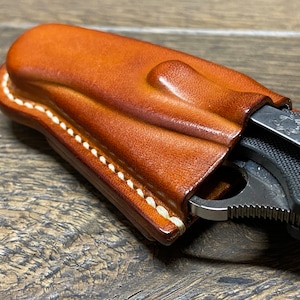 May include: A brown leather knife sheath with white stitching. The sheath is designed to hold a folding knife with a black handle and a silver blade.