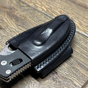 以下が含まれることがあります： 黒い革製のナイフシースに、ステンレス製の折りたたみナイフが部分的に挿入されています。ナイフはテクスチャード加工されたハンドルと銀色の刃を持っています。シースにはベルトクリップがあり、端に沿ってステッチが施されています。