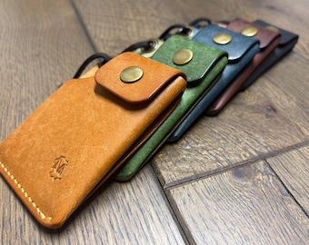 May include: A series of rectangular leather card holders arranged in a row, featuring colors including tan, green, blue, brown, and black. Each holder has a flap secured with a brass-toned snap button, a black cord loop, and visible stitching.