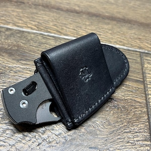 以下が含まれることがあります： 黒い革製のナイフシースに、一部挿入された折りたたみナイフ。シースには刻印されたエンブレムがあり、白い糸でステッチされています。ナイフは黒いハンドルと銀色の刃を持っています。アイテムは木製の表面にあります。