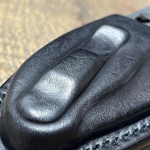 以下が含まれることがあります： 黒い革製のナイフシースのクローズアップ。シースは、テクスチャ加工された表面と、隆起した部分を備えた成形デザインを備えています。エッジはコントラストのある糸でステッチされており、ナイフの一部が見えます。