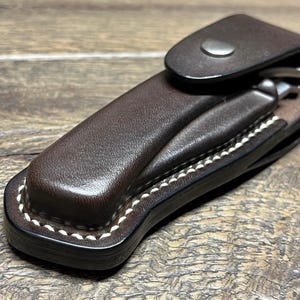 May include: A brown leather knife sheath with white stitching and a snap closure. The sheath is designed to hold a folding knife. The leather has a textured appearance.