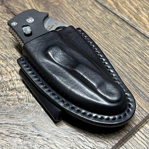 以下が含まれることがあります： 黒い革製のナイフシースに、一部挿入された折りたたみナイフ。シースは縁が縫い付けられており、成形されたデザインになっています。ナイフは黒いハンドルと銀色の刃を持っています。背景は木製の表面です。