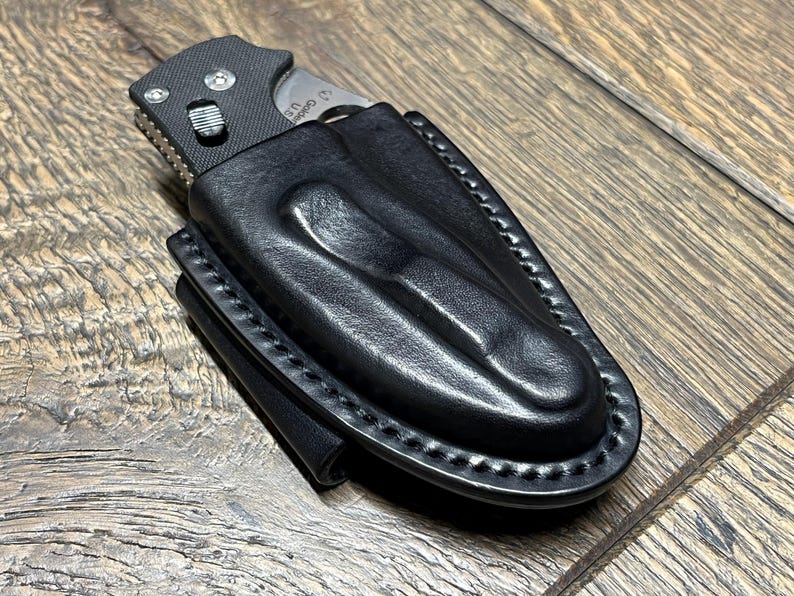 以下が含まれることがあります： 黒い革製のナイフシースに、一部挿入された折りたたみナイフ。シースは縁が縫い付けられており、成形されたデザインになっています。ナイフは黒いハンドルと銀色の刃を持っています。背景は木製の表面です。