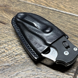 以下が含まれることがあります： 黒い革製のナイフシース。一部が見える折りたたみナイフ付き。ナイフは黒いハンドルと銀色の刃。シースはステッチされ、ベルトクリップが付いています。ナイフの刃には「Golden U.S.A.」の文字。