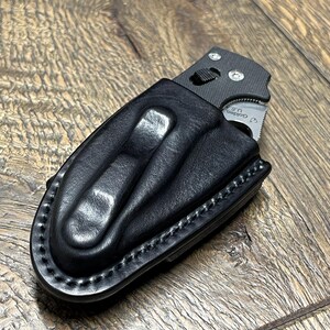以下が含まれることがあります： 黒い革製のナイフシースに、一部挿入された折りたたみナイフ。 シースにはベルトループがあり、縁に沿ってステッチが施されています。 ナイフの刃が見え、刃には「Golden U.S.」の文字があります。