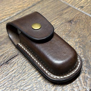 May include: A dark brown leather sheath with a flap closure secured by a brass-colored snap. The edges are stitched with white thread. The sheath is designed to hold a small tool or knife.