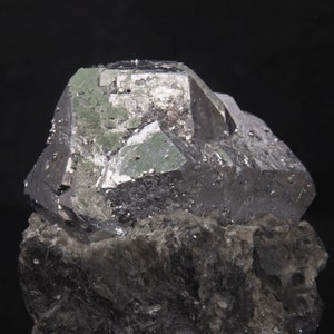 Può includere: Un grande esemplare minerale metallico grigio scuro con un rivestimento cristallino verde. Il minerale è posto su una roccia grigia ruvida.