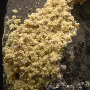 Può includere: Primo piano di un campione minerale giallo con una superficie ruvida e testurizzata. Il minerale è raggruppato in una forma densa e irregolare.
