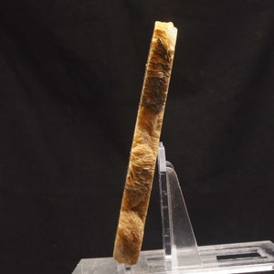 Op de afbeelding: Een rechthoekig mineraal specimen in lichtbruine tinten met donkerbruine insluitsels en een vezelachtige textuur. Het wordt gepresenteerd op een transparante acrylstandaard tegen een zwarte achtergrond. De schuine weergave toont het patroon.