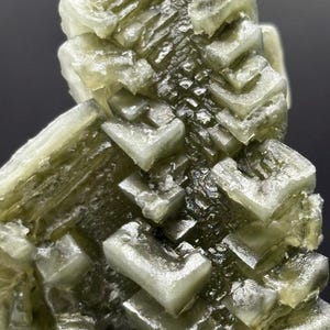 May include: Close-up of a mineral specimen with a crystalline structure. The mineral has a pale green hue and a translucent appearance. The crystals are blocky and stacked, creating a textured surface. The background is dark.