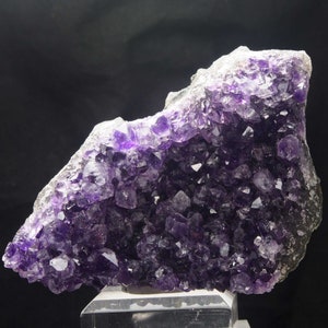 May include: A large amethyst geode in shades of deep purple. The crystal cluster is displayed on a clear stand, highlighting the natural formation and crystalline structure. The geode's interior is filled with sparkling, pointed crystals.