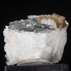 Może przedstawiać: Gromada białych kryształów kwarcu z dużym, ciemnoszarym, metalicznym okazym mineralnym osadzonym w środku. Okaz mineralny ma warstwową, prążkowaną strukturę.