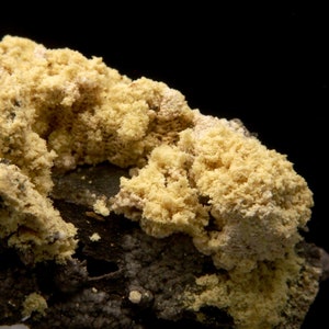 Può includere: Primo piano di un campione minerale giallo con una superficie ruvida e testurizzata. Il minerale sembra crescere su una roccia grigio scuro.