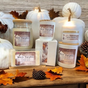 May include: A collection of white pumpkin scented candles in various sizes. The candles are in glass jars with wooden lids and feature a label that reads "White Pumpkin" with the scent description "whipped vanilla pumpkin spice".