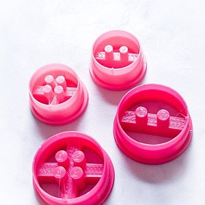 May include: Four pink plastic cookie cutters with different designs. Each cutter has a raised design in the center. The cutters are on a white surface.