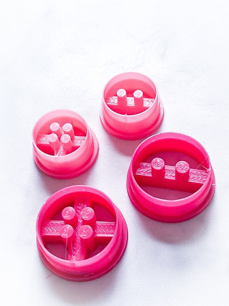 May include: Four pink plastic cookie cutters with different designs. Each cutter has a raised design in the center. The cutters are on a white surface.
