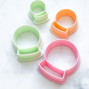 May include: Set of four plastic cookie cutters shaped like snow globes. The cutters are in different sizes and colours: green, pink, and orange.