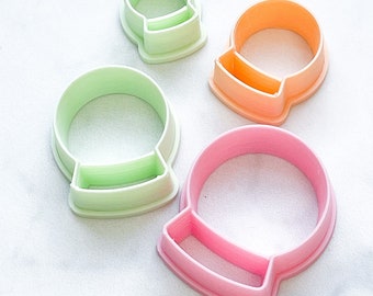 May include: Set of four plastic cookie cutters shaped like snow globes. The cutters are in different sizes and colors: green, pink, and orange.