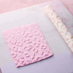 May include: A white plastic rolling pin with a leopard print design is shown next to a pink fondant sheet with the same design embossed into it.