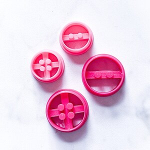 May include: Four pink plastic cookie cutters shaped like Lego bricks. The cutters are different sizes and have different numbers of studs.