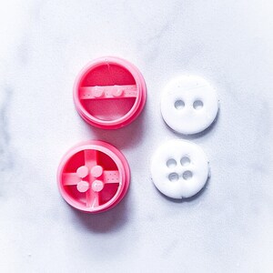 May include: Two pink plastic button molds and two white plastic buttons. The molds have four holes in the center and the buttons have two and four holes.