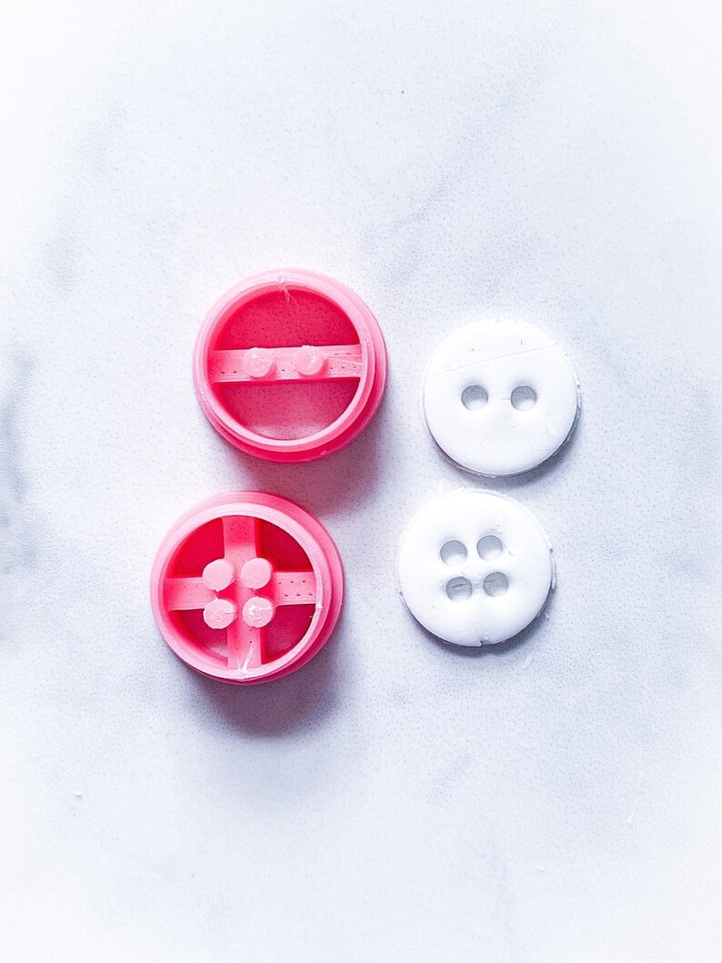 May include: Two pink plastic button molds and two white plastic buttons. The molds have four holes in the center and the buttons have two and four holes.