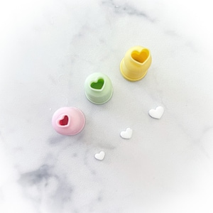 May include: Three silicone piping tips with heart-shaped openings. The tips are yellow, green, and pink. Four white heart-shaped sprinkles are scattered around the tips.