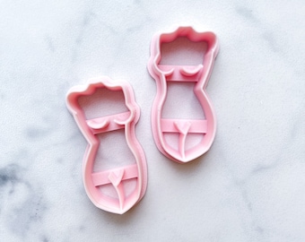 May include: Two pink plastic cookie cutters shaped like a female figure wearing a bikini.