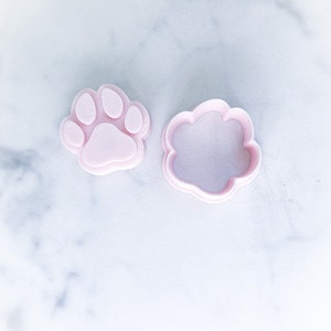 May include: Two light pink paw print-shaped cookie cutters. One is a solid paw print, and the other is the outline of a paw print. The cutters are plastic and are on a white marble surface.