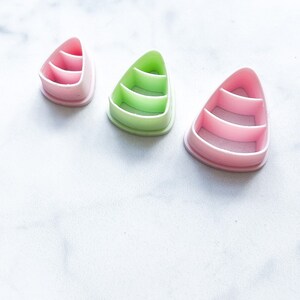 Puede incluir: Tres cortadores de galletas de plástico con forma de triángulo con tres líneas horizontales en el interior. Los cortadores son de color rosa y verde.