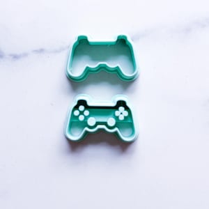 May include: Two teal-colored cookie cutters in the shape of a video game controller. The top cutter is a solid outline, while the bottom one has detailed button and directional pad cutouts. Ideal for baking themed treats.