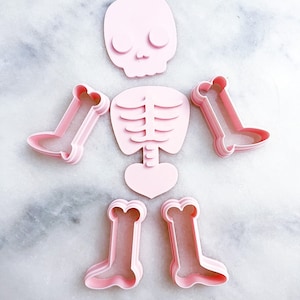 Puede incluir: Cortadores de galletas de esqueleto de plástico rosa. El juego incluye un cráneo, una caja torácica y dos pares de piernas.