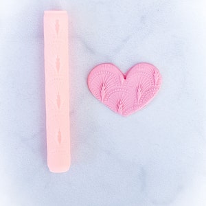 以下が含まれることがあります： 装飾的なパターンが施されたピンクのシリコン製麺棒と、同様のパターンが施されたピンクのハート型のフォンダンカッターです。