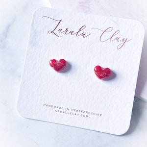 Puede incluir: Un par de pequeños pendientes en forma de corazón rojo hechos de arcilla polimérica. Los pendientes están hechos a mano en Hertfordshire, Inglaterra. El texto "LaraLu Clay" está impreso en la tarjeta.