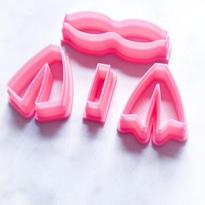May include: A set of five pink plastic cookie cutters shaped like a bow, two separate bow pieces, and a rectangle.