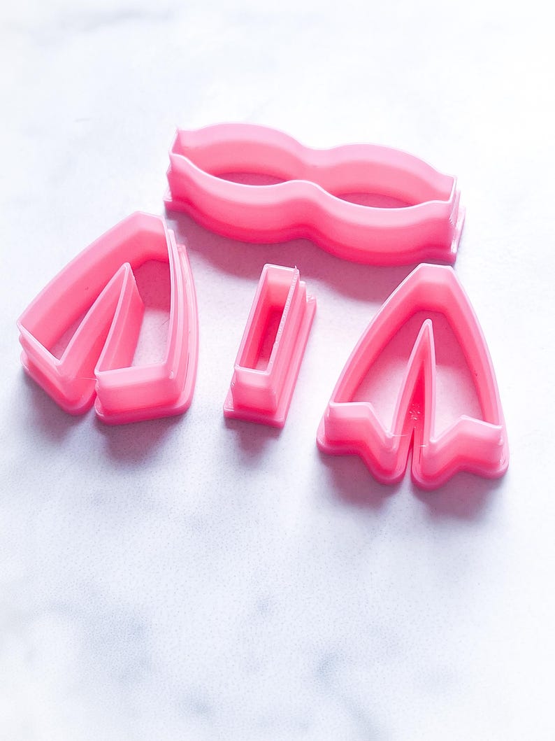 May include: A set of five pink plastic cookie cutters shaped like a bow, two separate bow pieces, and a rectangle.