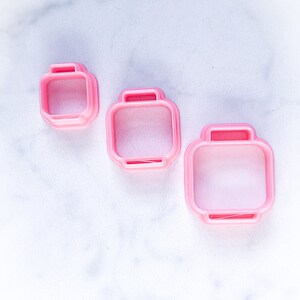 May include: Three pink plastic cookie cutters shaped like Lego bricks. The cutters are different sizes.